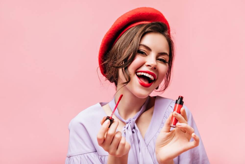 adorable-curly-woman-paints-her-lips-with-red-lipstick-laughs-pink-background
